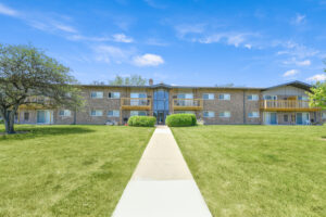Exterior brick Residential buildings, large field of grass, Sidewalk leading to building, meticulous landscaping, lush foliage, photo taken a sunny day.