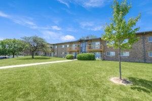 Exterior brick Residential buildings, large field of grass, Sidewalk leading to building, meticulous landscaping, lush foliage, photo taken a sunny day.