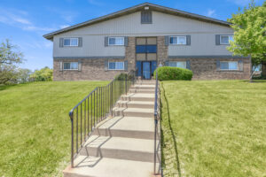 Exterior brick Residential buildings, large field of grass, Sidewalk steps leading to building, meticulous landscaping, lush foliage, photo taken a sunny day.