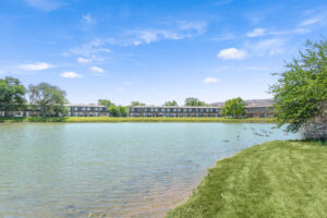 Exterior Body of water at Acclaim at Hinsdale Lake, Ducks swimming, residential buildings in the distance, meticulous landscaping, lush foliage, photo taken on a sunny day.