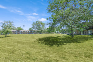 Exterior Large grass field, large tree with lush foliage, meticulous landscaping, brick residential buildings in the distance, photo taken on a sunny day.