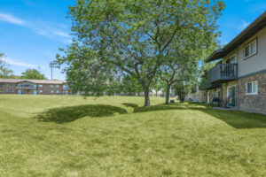 Exterior Large grass field, large tree with lush foliage, meticulous landscaping, brick residential buildings in the distance, photo taken on a sunny day.
