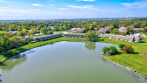 Aerial Exterior of Acclaim at Hinsdale Lake's body of water, brick residential buildings in the background, lush foliage, meticulous landscaping, photo taken on a sunny day.