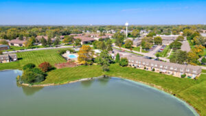 Aerial Exterior Acclaim at hinsdale lake, body of water multiple brick residential buildings, lush foliage, meticulously landscaped property, tree line in the distance, photo taken on a sunny day.