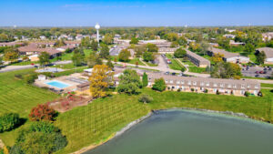 Aerial Exterior Acclaim at hinsdale lake, body of water, multiple brick residential buildings, community pool, playground, lush foliage, meticulously landscaped property, tree line in the distance, photo taken on a sunny day.