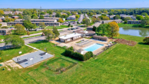Aerial Exterior of Community Amenity building, Large sport court, full length swimming pool, large lounge chaise area, playground lush foliage, Meticulously groomed grounds, photo taken on a sunny day.