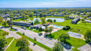Aerial Exterior of Community amenity building, large pool, meticulous landscaping, lush foliage, body of water in the background, treeline in the distance, photo taken on a sunny day.