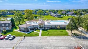 Aerial Exterior of Community amenity building, large pool, meticulous landscaping, lush foliage, body of water in the background, treeline in the distance, photo taken on a sunny day.