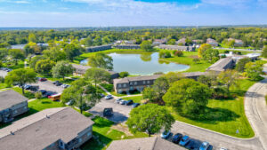 Aerial Exterior of brick residential buildings, lush foliage, meticulously groomed grounds, treeline in the distance, photo taken on a sunny day.