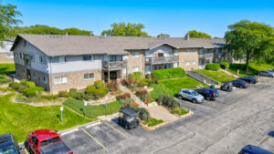 Aerial Exterior of Brick Residential Buildings, meticulously groomed grounds, sidewalks leading to buildings, lush foliage, treeline in the distance, photo taken on a sunny day.