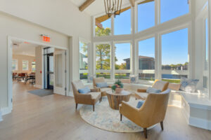 Interior clubhouse, contemporary rustic decor, high ceilings, large bay window with bench seating and 4 lounge chairs around a rustic coffee table, wood floors.