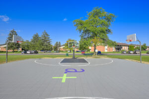Exterior Sport court, 6 basketball hoops, avanath logo center court, meticulous groomed grounds around the court, lush foliage, residential buildings in the background, photo taken on a sunny day.