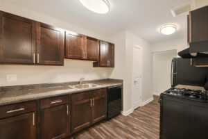 Interior unit kitchen, stone countertops, brown cabinetry, black appliances, stainless steel sink, wood floors, neutral toned walls.