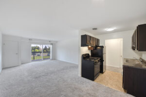 Interior Unit Living room/ kitchen, open floor plan, black appliances in kitchen, stainless steel dual sink, tile floor in kitchen area, neutral toned carpeting in living room, white walls, sliding glass doors to balcony area.